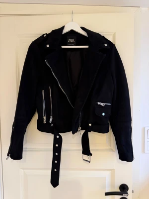 Svart mockajacka från Zara - Snygg svart mockajacka från Zara med klassisk bikerstil. Jackan har silvriga dragkedjor, bälte i midjan och flera fickor med detaljer. Croppad modell med långa ärmar.