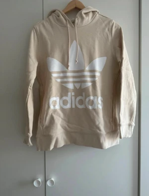 Beige Adidas hoodie med logga - Säljer en beige hoodie från Adidas med det klassiska trefoil-loggan i vitt framtill. Tröjan har huva med snörning och långa ärmar. Perfekt för både vardag och chill dagar