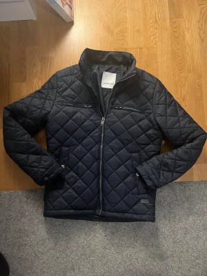 Mörkblå quiltad jacka Lindbergh - Quiltad mörkblå jacka från Lindbergh med snyggt rutmönster, två bröstfickor med dragkedja och ståkrage. Jackan har hel dragkedja framtill och är tillverkad i lätt polyester. Perfekt för dig som gillar stilren och modern look. Storleken är S och den är helt ny och inga defekter 