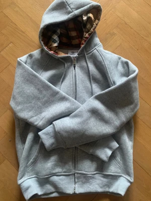 Grå hoodie från Burberry med rutig huva - Snygg grå hoodie från Burberry i storlek M. Utsidan är mjuk och har en klassisk dragkedja med Burberry-logga. Insidan av huvan har det ikoniska rutiga Burberry-mönstret i beige, svart och rött. Perfekt för dig som gillar stilrena detaljer och exklusiva märken. Pris kan diskuteras. Om du är intresserad av att köpa så så ska du veta att jag inte kan skicka mellan 17 December och 5 Januari