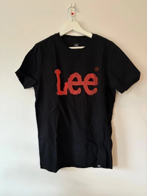 Svart Lee t-shirt med 3D-logga - Svart t-shirt från Lee i bomull med coolt 3D-textur på den röda loggan framtill. Regular fit och klassisk rund hals. Kortärmad modell med Lee-logga även i nacken. Perfekt för dig som gillar streetwear och vill sticka ut med en unik detalj.