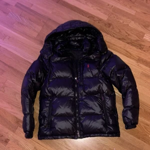 Svart pufferjacka från Polo Ralph Lauren - Snygg svart pufferjacka från Polo Ralph Lauren med huva och dragkedja. Jackan har en glansig finish, två sidofickor med dragkedja och klassisk röd logga på bröstet. Perfekt för kalla dagar när du vill hålla stilen.