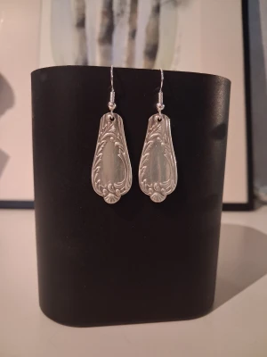 Handgjorda örhängen återbruk  - Egentillverkade örhängen skapade av 2st bestick gjorda i nysilver. Örhänget består av olika typer av snirklar längs kanterna och blir en stilren och elegant accessoar till outfiten. Längd ca 3.4cm. Öronkroken är nickelsäker och aldrig använd.
