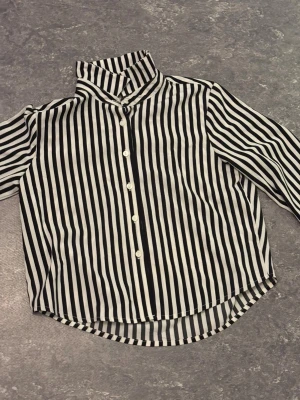 Svartvit randig skjorta med knappar - Snygg skjorta med svartvita vertikala ränder och klassisk krage. Skjortan har långa ärmar och vita knappar framtill. Materialet känns lätt och luftigt, perfekt för dig som gillar stilrena och grafiska plagg.