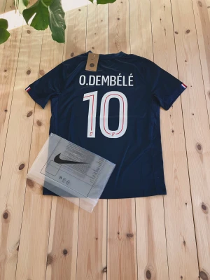 PSG tröja O. Dembélé #10 Nike - Snygg marinblå Paris Saint-Germain tröja med O. Dembélé och nummer 10 på ryggen. Tröjan har vita och röda detaljer och är tillverkad i lätt polyester, perfekt för fotbollsfans. Officiell Nike-produkt med PSG-märke.