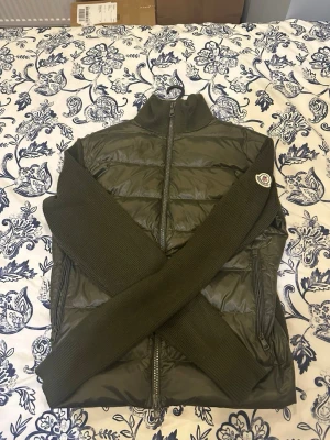 Olivgrön pufferjacka från Moncler - Snygg olivgrön pufferjacka från Moncler med stickade ärmar och hög krage. Jackan har dragkedja framtill och sidofickor med dragkedja. Moncler-logga på ärmen och quiltad front i syntetmaterial. Perfekt för dig som vill ha en clean och modern look.