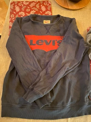 Svart Levi's sweatshirt med röd logga - Cool svart sweatshirt från Levi's med stor röd logga framtill. Tröjan har rund hals, långa ärmar och ribbade muddar vid ärmslut och nederkant. Perfekt för dig som gillar klassisk streetstyle och vill ha en bekväm tröja med snygg branding. Storleken är 16 år, ungefär som en Smal 