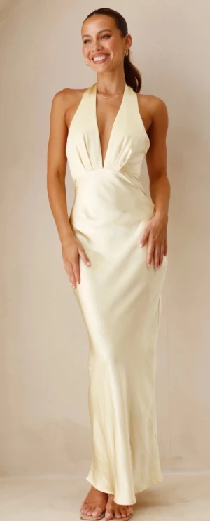 Champagnefärgad långklänning i satin - Säljer en elegant champagnefärgad långklänning med djup v-ringning och halterneck. Klänningen har en slimmad passform och är i satinmaterial med öppen rygg 