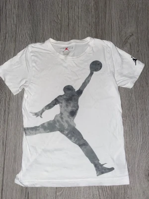 Vit Jordan t-shirt med stor logga - Snygg vit t-shirt från Jordan i bomull med stor Jumpman-tryck i grått på framsidan och mindre logga på ärmen. Klassisk rund hals och korta ärmar, perfekt för dig som gillar sportig streetstyle. Passar dig som vill ha en clean look med ikonisk basketstil.