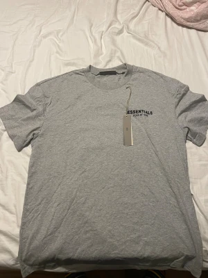 Grå Essentials Fear of God t-shirt - Snygg grå t-shirt från Essentials Fear of God med svart logotyptryck både fram och bak. Klassisk rund hals och korta ärmar. Mjuk och skön bomullskvalitet som passar perfekt till streetwear-stilen. Diskret svart patch med logga i nacken.