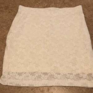 Vit spetskjol med blommönster - Söt vit kortkjol med blommig spets och diskret mönster. Kjolen har en rak passform och är perfekt för dig som gillar en romantisk och feminin stil. Spetsdetaljerna ger kjolen en extra touch av elegans.