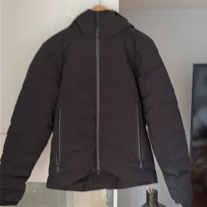 Svart pufferjacka från Sail Racing - Svart pufferjacka från Sail Racing i storlek medium. Jackan har en stilren design med raka linjer, huva och tre dragkedjor framtill. Materialet är syntet och jackan har ett diskret Sail Racing-logomärke på ärmen. Perfekt för kalla dagar och snygg till streetwear.