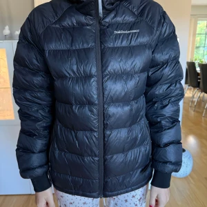 Svart dunjacka Peak Performance S - Svart dunjacka från Peak Performance i storlek S. Jackan har quiltad design, lång ärm och dragkedja framtill. Broderad logga på bröstet och ärmen. Perfekt för kalla dagar, snygg och enkel att matcha med din stil. Jackan har en liten defekt fram till på höger sida! 