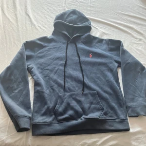 Mörkblå hoodie från Polo Ralph Lauren - Snygg mörkblå hoodie från Polo Ralph Lauren med klassisk röd broderad logga på bröstet. Tröjan har huva med svarta snören, stor magficka och långa ärmar. Perfekt för chill dagar och enkel att matcha med jeans eller joggers.