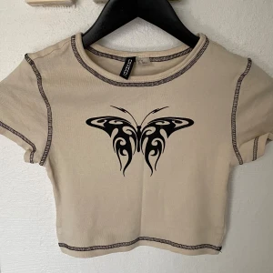 Beige croppad topp med tribal fjäril - Trendig croppad topp från H&M i beige med kontrastsömmar och svart tribal fjäril-tryck framtill. Kortärmad modell med rund halsringning och figurnära passform. Perfekt för dig som gillar edgy och unik stil.
