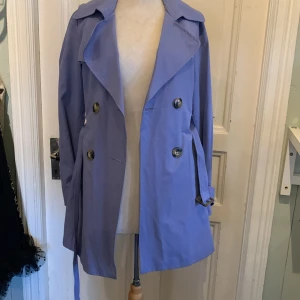  trenchcoat från Benetton  - Snygg trenchcoat i syren från United Colors of Benetton med klassisk dubbelknäppning, axelklaffar och bälte i midjan. Jackan har långa ärmar och stora knappar framtill. Perfekt för dig som vill sticka ut med färg i vår eller höst.