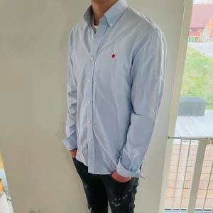 Ljusblå skjorta från Morris - Snygg ljusblå skjorta från Morris med klassisk button-down krage och vit knappstängning. Skjortan har en röd broderad logga på bröstet och är tillverkad i bomull. Perfekt för dig som gillar stilrena och tidlösa plagg. Storlek 40 vilket passar mellan Medium och Large. Snabb Leverans!