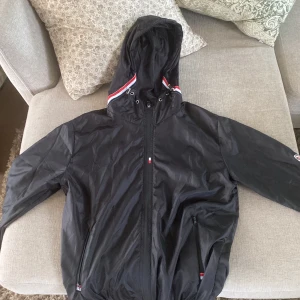 Svart vindjacka från Moncler med huva - Svart vindjacka från Moncler med huva och dragkedja framtill. Jackan har Moncler-logga på ärmen, röda och vita detaljer vid huvan och två fickor med dragkedja. Tillverkad i lätt polyester, perfekt för blåsiga dagar.