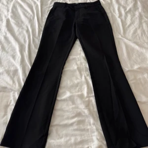 Svarta bootcut kostymbyxor BikBok - Svarta kostymbyxor från BikBok i storlek 36. Byxorna har bootcut-modell med pressveck, bälteshällor och stängs med dragkedja och knapp. Snygg passform och klassisk look, perfekta till en trendig outfit.