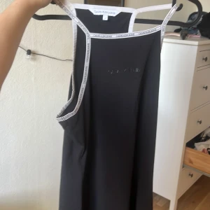 Svart ärmlös klänning Calvin Klein Jeans - Svart ärmlös klänning från Calvin Klein Jeans i mjuk bomull. Klänningen har vita kantband med logotyp längs axelbanden och runt halsen. Enkel och sportig design med broderad logga framtill. Perfekt för en avslappnad och trendig look.