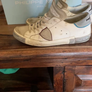 Philippe Model vita sneakers med blå detalj - Säljer ett par vita Philippe Model sneakers med grå och blå detaljer. Skorna har snörning, rund tå och platt sula. Märkets ikoniska sköld-logga syns på sidan och materialet är skinn med inslag av mocka. Perfekta för dig som gillar en clean men ändå unik look.