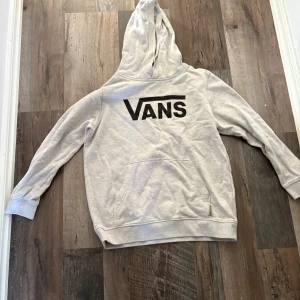 Ljusgrå hoodie från Vans - Säljer en ljusgrå hoodie från Vans med stor svart logga på bröstet. Tröjan har huva, magficka och långa ärmar. Perfekt för dig som gillar streetwear och vill ha en enkel men snygg look.