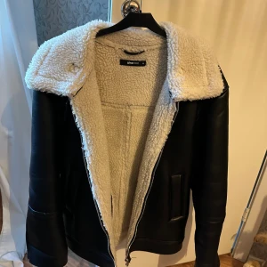 Svart teddyjacka från Gina Tricot XS - Säljer en svart teddyjacka från Gina Tricot i storlek XS. Jackan har mjukt vitt teddyfoder, bred krage, dragkedja och snyggt spänne vid halsen. Utsidan är i skinnimitation och insidan är fluffig och varm. Perfekt för kalla dagar!