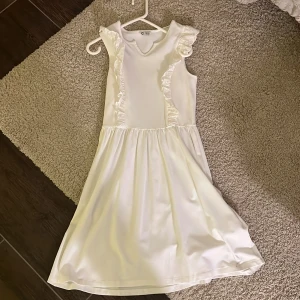 Vit ärmlös klänning med volang - Superfin vit klänning i ärmlös modell med volangdetaljer längs axlarna och bröstet. Klänningen har en mjuk och skön, är i barnstorlek men stretchiga så passar xs och s också. Nästan aldrig använt.