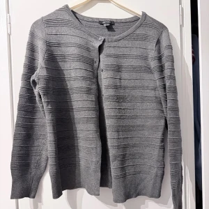 Grå ribbad kofta från Esmara - Grå kofta från Esmara med ribbade horisontella ränder och rund halsringning. Långärmad modell med knäppning framtill och diskreta knappar. Perfekt för lager-på-lager och enkel att matcha med andra plagg. Men som man kan se på bild saknas det en knapp men inget man lägger märke till 