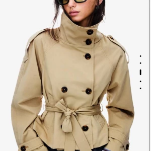 Kort beige trenchcoat med bälte - Snygg kort trenchcoat i beige med dubbelknäppning, stora knappar och bälte i midjan. Jackan har axelklaffar, hög krage och långa ärmar med knäppning vid ärmslut. Från bershka helt slutsåld 