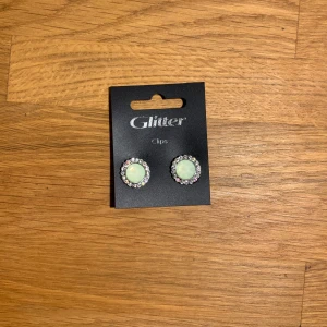 Mintgröna clipsörhängen från Glitter - Runda clipsörhängen med mintgrön sten i mitten, omgiven av små gnistrande stenar i silverfärg. Klassisk och söt design som ger en glittrig touch till din look. Perfekta för dig som vill ha örhängen utan hål. Ena örhängets gummi har hårdnat men funkar lika bra 
