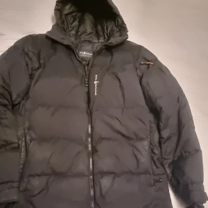 Svart jacka från Sail Racing - Svart pufferjacka från Sail Racing med huva och dragkedja framtill. Jackan har en broderad logga på huvan och en diskret logga på bröstet. Se bilder. 