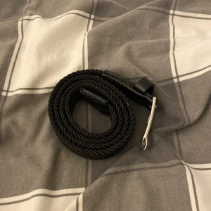 Svart flätat bälte från Kappahl - Snyggt svart flätat bälte från Kappahl med silverfärgat spänne. Bältet är gjort i ett mjukt syntetmaterial med detaljer i skinnimitation. Perfekt accessoar för att lyfta din outfit med en stilren touch.