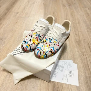 Maison Margielas  - Maison Margiela sneakers i vitt skinn och mocka med unikt paint splatter-mönster i blått, rött, gult och svart. Klassisk låg modell med snörning och brun gummisula. Perfekt för dig som vill sticka ut med en konstnärlig vibe.