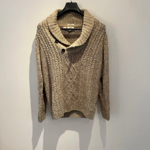 Beige stickad tröja från PL&CO - Mysig beige stickad tröja från PL&CO med grov kabelstickning och bred krage med två knappar. Tröjan har lång ärm och ribbade muddar, perfekt för kyliga dagar. Den är använd men inga defekter, hör av er vid fler bilder eller frågor