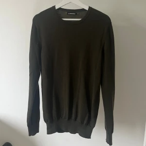 Mörkgrön stickad tröja från J.Lindeberg - En stilren mörkgrön stickad tröja från J.Lindeberg med rund halsringning och diskret logga på bröstet. Tröjan har lång ärm och ribbade muddar vid ärmslut och nederkant. Perfekt för lager-på-lager och snygg till jeans.