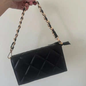 Svart quiltad axelväska med kedjedetalj - Snygg svart axelväska i quiltad design med guldfärgad kedja flätad med skinn och justerbar axelrem. Väskan har dragkedja upptill och stilren, rektangulär form. Perfekt accessoar för att lyfta din outfit.