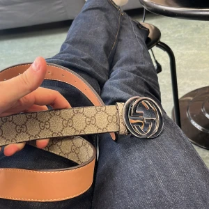 Beige bälte - Säljer ett snyggt beige bälte med klassiskt mönster och stor silverfärgad logospänne. Bältet är gjort i skinn och har en stilren form med bruna och beige toner. Perfekt för dig som vill ha en ikonisk accessoar med lyxig känsla.   Köpte av en kompis.  Pris kan diskuteras😀