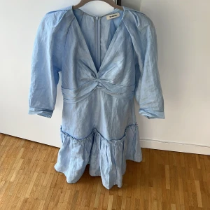 Ljusblå kortklänning från Sandro - Supersöt ljusblå klänning från Sandro med v-ringning, twistad detalj framtill och volangkant nertill. Klänningen har trekvartsärmar och dragkedja i ryggen. Tillverkad i ett lätt, glansigt material som ger en lyxig känsla.