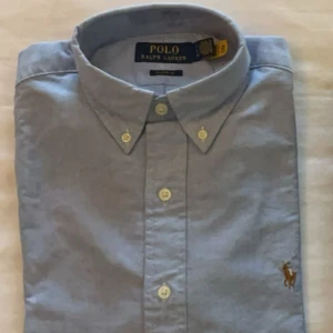 Ljusblå skjorta från Polo Ralph Lauren - Helt ny aldrig använd! Polo ralph lauren sjorta, skick 10/10. Färgen kan uppskattas som lite grå aktig men är bara dåligt ljus på bilden, (den har inga missfärgningar) den är den klassiska ljus blå Ralph lauren sjort färgen! Priset är ej hugget i sten! Nypris 1600. Om du har några frågor eller funderingar, så är det bara att höra av sig!