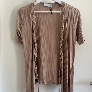 Beige ribbad bolerotopp från Three Sixty Five - Säljer en beige bolerotopp från Three Sixty Five i storlek small. Toppen har korta ärmar, ribbad struktur och volangdetaljer längs öppningen framtill. Materialet är mjukt och lätt, perfekt för lager-på-lager. 