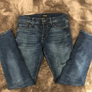 Blå jeans från Jack & Jones - Säljer ett par snygga slim fit blå jeans från Jack & Jones med klassisk femficksdesign och diskreta slitningar. Jeansen har normal passform och raka ben, med kontrastsömmar och knappgylf. Perfekta för dig som gillar stilrena och bekväma jeans.