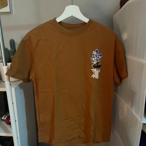 Brun t-shirt från H&M - Säljer en brun t-shirt från H&M med relaxed fit. Framsidan har ett litet tryck med blå blommor och texten 'Talk to me', medan baksidan har ett större blommotiv och samma text. Skön bomullskvalitet och korta ärmar, perfekt för en chill stil.