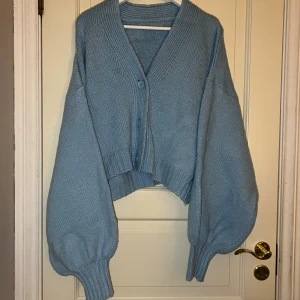 Ljusblå stickad kofta med ballongärmar - Superfin ljusblå stickad kofta med oversized ballongärmar och bred ribbad mudd. Koftan har v-ringning, knappar framtill och croppad passform. Perfekt att slänga över en topp. Den är i storlek M men är oversized. Det finns även fläckar som syns på bilden.