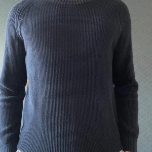 Mörkblå stickad tröja från East West - Mörkblå stickad tröja från East West i storlek M. Tröjan har en klassisk rund halsringning och är ribbstickad vid hals, ärmslut och nederkant. Perfekt för kyligare dagar och enkel att matcha med jeans eller chinos.