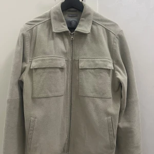 Beige mockajacka Human Scales Rick S - Snygg beige mockajacka från Human Scales, modell Rick. Jackan har två stora bröstfickor med lock, raka sidfickor och dragkedja framtill. Klassisk krage och knappar vid ärmslut. Perfekt för dig som gillar stilren och clean look.