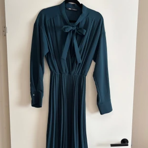 Plisserad mörkgrön långklänning från Zara - Säljer en elegant mörkgrön långklänning från Zara med plisserad kjol, långärmade ärmar och snygg knytrosett vid halsen. Klänningen har markerad midja och breda manschetter med knappar. Perfekt för dig som gillar stilrena och klassiska plagg. Endast använd 1 gång. Nyskick. 