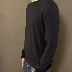 Svart långärmad tröja i bomull - En stilren svart långärmad tröja i mjuk bomull. Tröjan har rund halsringning och en klassisk passform som funkar till jeans eller andra byxor. Perfekt för dig som gillar enkel och clean stil.