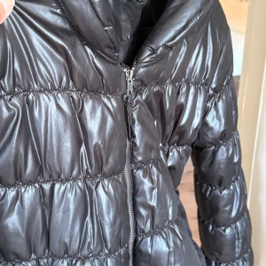 Svart pufferjacka från Divided - Säljer en svart pufferjacka från Divided med glansig finish och huva. Jackan har dragkedja framtill och två fickor med dragkedja. Perfekt för kyliga dagar och har en riktigt skön, puffig känsla.