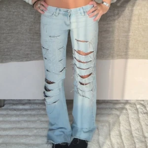 Ljusblå slitna jeans med tryck - Säljer ett par ljusblå jeans med rejält slitna detaljer och stora hål framtill. Jeansen har ett tryck på låret och klassisk femficksmodell. Modellen är utsvängd med låg midja och tillverkad i jeansmaterial. Perfekta för dig som gillar en edgy streetstil. Säljer då dom aldrig andvänts!!🫰🏼❤️❤️